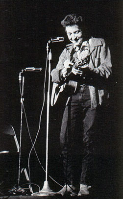 Black-and-white photo of a young Bob Dylan performing with an acoustic guitar at two microphone stands